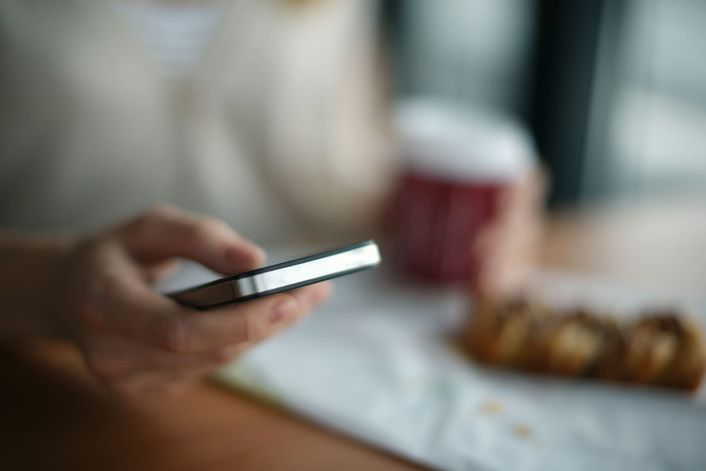 Een hand met een telefoon aan tafel, met een beker koffie en gebak op de achtergrond.