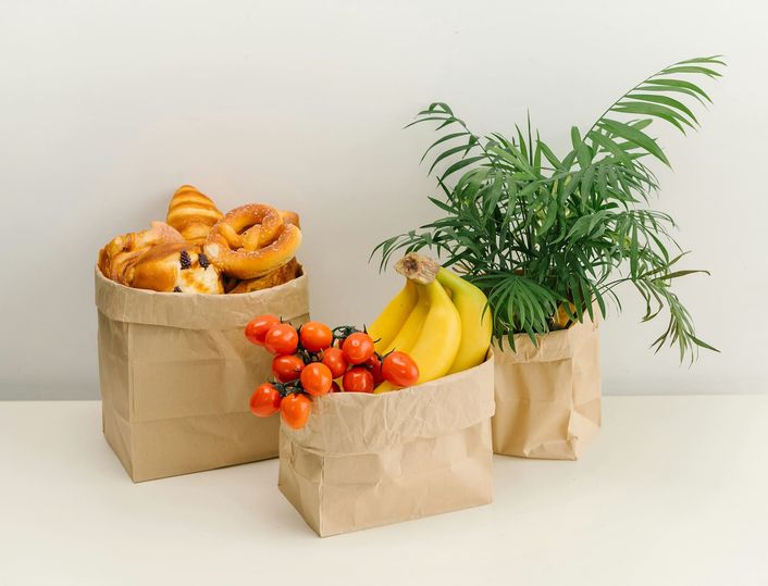 Drie papieren tassen met brood, tomaten, bananen en een plant erin.